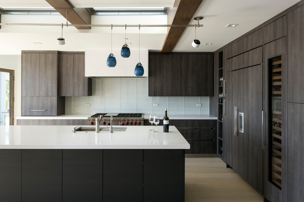 Design ideas for a large modern l-shaped open plan kitchen in San Diego with an undermount sink, flat-panel cabinets, dark wood cabinets, quartz benchtops, white splashback, glass tile splashback, panelled appliances, light hardwood floors, with island and white benchtop.