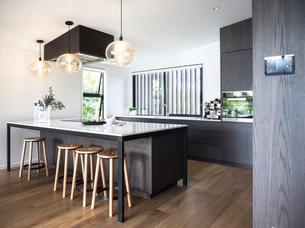 Photo of a mid-sized contemporary l-shaped eat-in kitchen in Auckland with an undermount sink, flat-panel cabinets, dark wood cabinets, quartz benchtops, with island, white splashback, medium hardwood floors, brown floor and white benchtop.