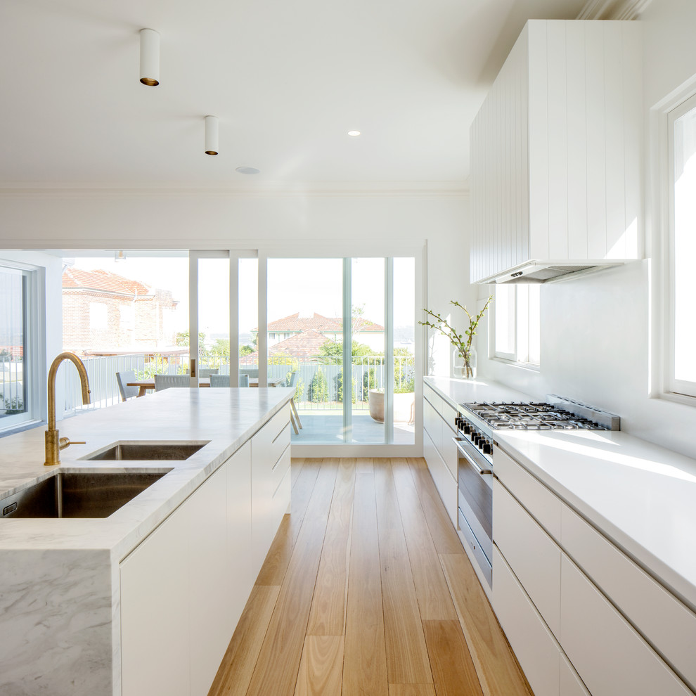 Photo of a modern galley kitchen in Sydney with an undermount sink, flat-panel cabinets, white cabinets, white splashback, stainless steel appliances, light hardwood floors, with island, beige floor and white benchtop.