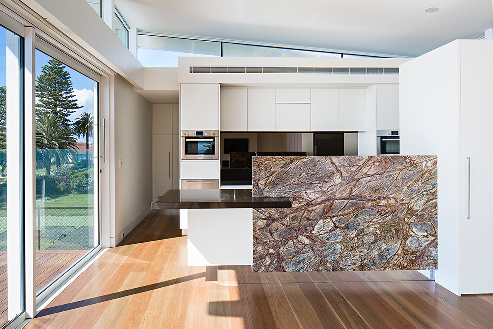 Design ideas for a mid-sized contemporary galley open plan kitchen in Sydney with an undermount sink, flat-panel cabinets, white cabinets, quartz benchtops, metallic splashback, glass sheet splashback, stainless steel appliances, medium hardwood floors and with island.
