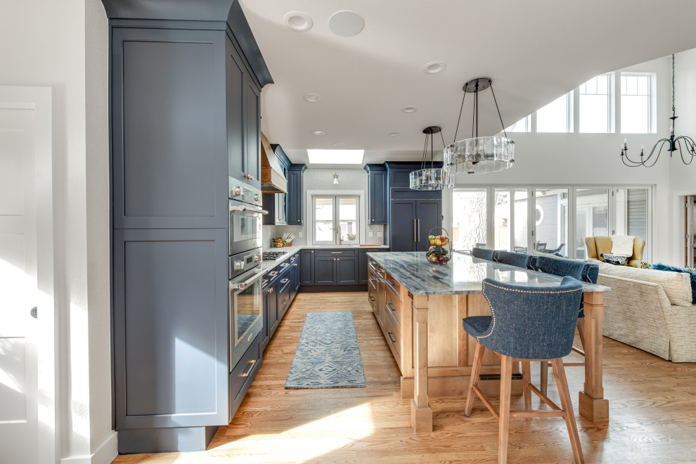 Inredning av ett klassiskt stort blå blått kök, med en undermonterad diskho, luckor med infälld panel, blå skåp, bänkskiva i kvartsit, vitt stänkskydd, stänkskydd i keramik, integrerade vitvaror, ljust trägolv, en köksö och brunt golv
