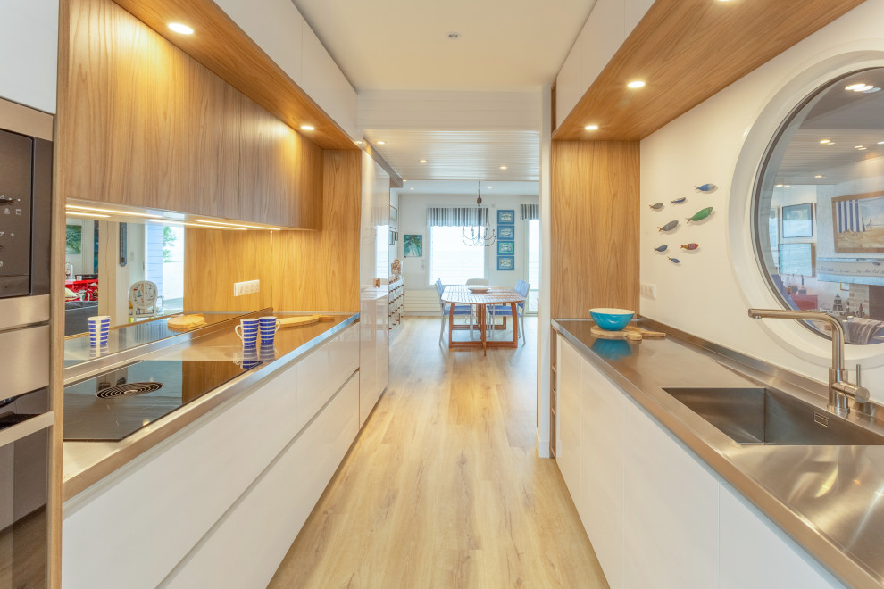 Design ideas for a small beach style galley open plan kitchen in Rennes with an integrated sink, beaded inset cabinets, white cabinets, stainless steel benchtops, mirror splashback, stainless steel appliances, light hardwood floors and timber.
