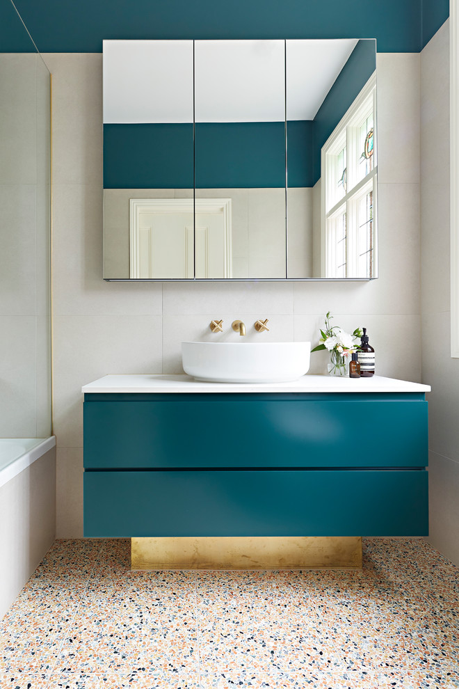 Small contemporary master bathroom in Melbourne with beige tile, beige walls, terrazzo floors, a vessel sink, multi-coloured floor, white benchtops, green cabinets, a shower/bathtub combo, a hinged shower door, a drop-in tub, a wall-mount toilet, stone tile, engineered quartz benchtops and flat-panel cabinets.