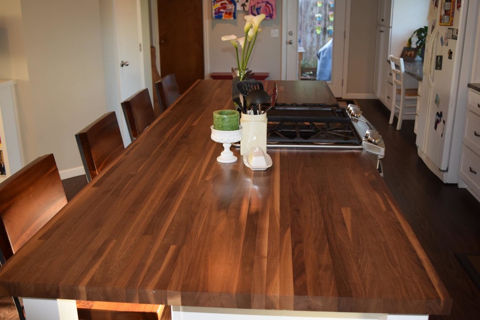 Example of a mid-sized transitional galley dark wood floor and brown floor open concept kitchen design with an undermount sink, shaker cabinets, white cabinets, wood countertops, white backsplash, porcelain backsplash, white appliances and an island