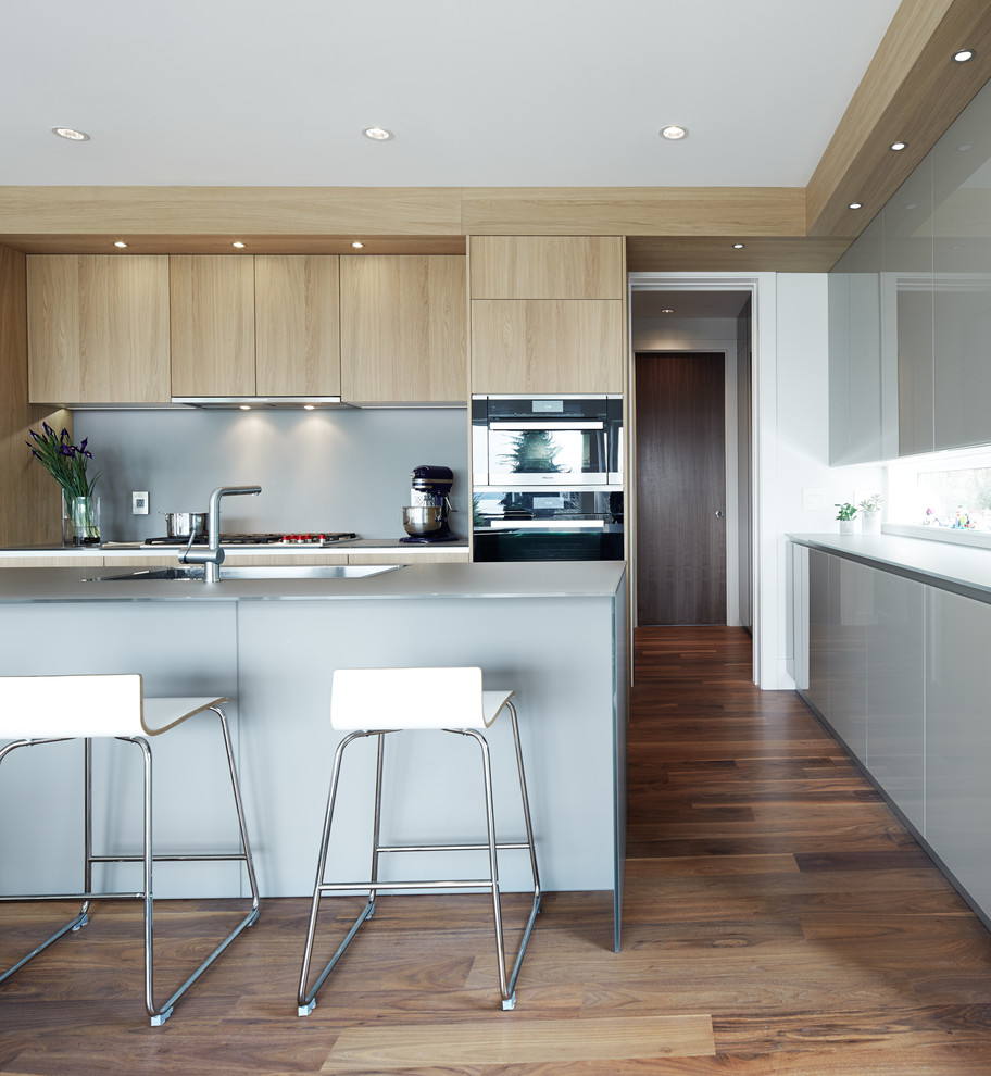 This is an example of a mid-sized contemporary l-shaped open plan kitchen in Vancouver with an undermount sink, flat-panel cabinets, light wood cabinets, glass benchtops, grey splashback, stainless steel appliances, dark hardwood floors and with island.