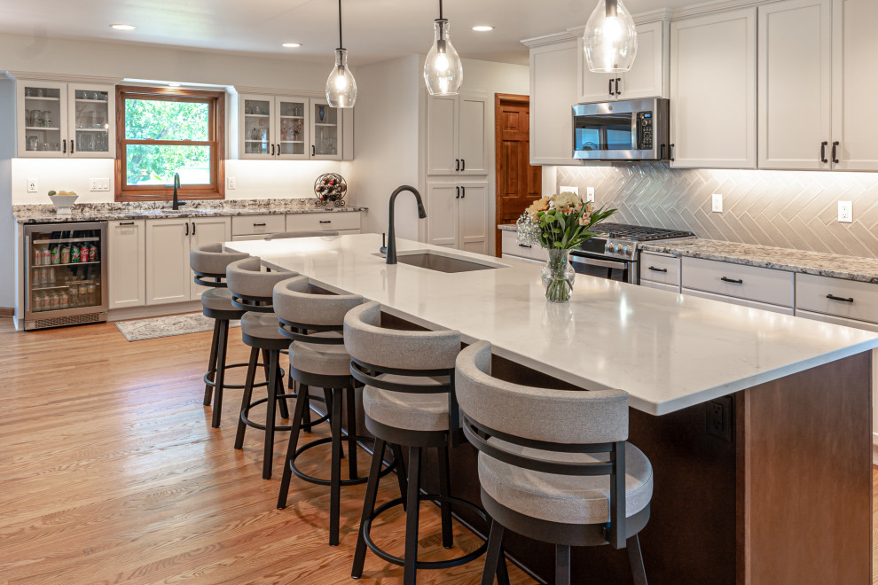 Photo of a large transitional l-shaped eat-in kitchen in Milwaukee with an undermount sink, recessed-panel cabinets, white cabinets, granite benchtops, grey splashback, mosaic tile splashback, stainless steel appliances, medium hardwood floors, with island, brown floor and multi-coloured benchtop.