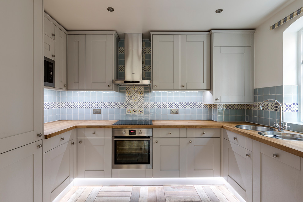 Design ideas for a mid-sized traditional u-shaped eat-in kitchen in London with a double-bowl sink, raised-panel cabinets, grey cabinets, wood benchtops, blue splashback, mosaic tile splashback, black appliances, cement tiles and no island.