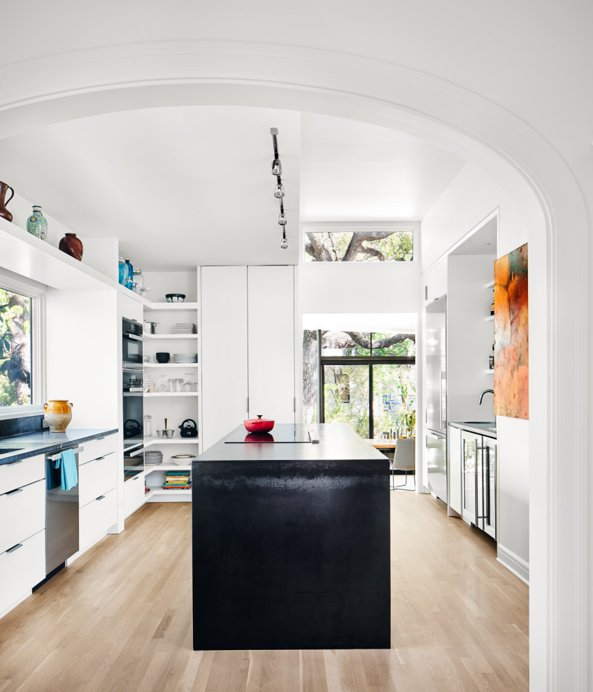 This is an example of a contemporary u-shaped kitchen in Austin with an undermount sink, flat-panel cabinets, white cabinets, stainless steel appliances, light hardwood floors, with island, beige floor and black benchtop.