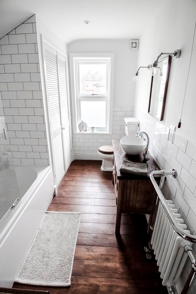 Mid-sized traditional kids bathroom in London with shaker cabinets, brown cabinets, a drop-in tub, a shower/bathtub combo, a one-piece toilet, white tile, subway tile, white walls, dark hardwood floors, a vessel sink, wood benchtops, brown floor and a sliding shower screen.