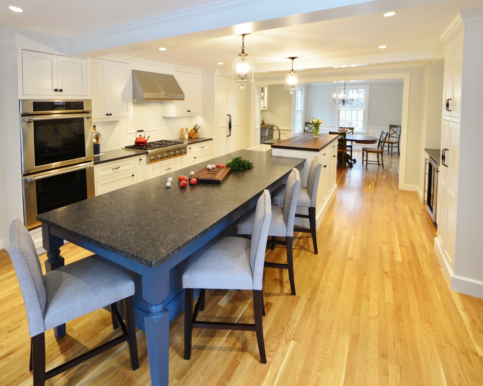 Photo of a large traditional eat-in kitchen in Boston with a farmhouse sink, flat-panel cabinets, white cabinets, granite benchtops, white splashback, ceramic splashback, stainless steel appliances, light hardwood floors and with island.
