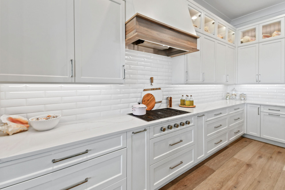 This is an example of a mid-sized beach style l-shaped kitchen in Other with white cabinets, quartz benchtops, white splashback, subway tile splashback, panelled appliances, light hardwood floors, with island, brown floor, white benchtop and recessed.