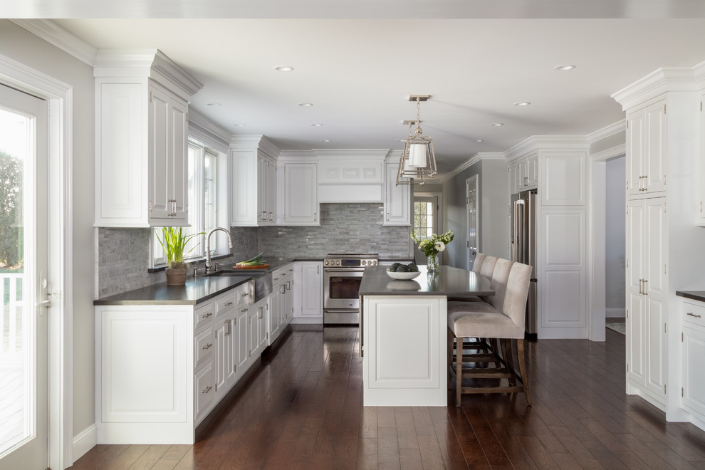Traditional u-shaped kitchen in New York with a farmhouse sink, raised-panel cabinets, white cabinets, grey splashback, stainless steel appliances, dark hardwood floors, with island, brown floor and grey benchtop.