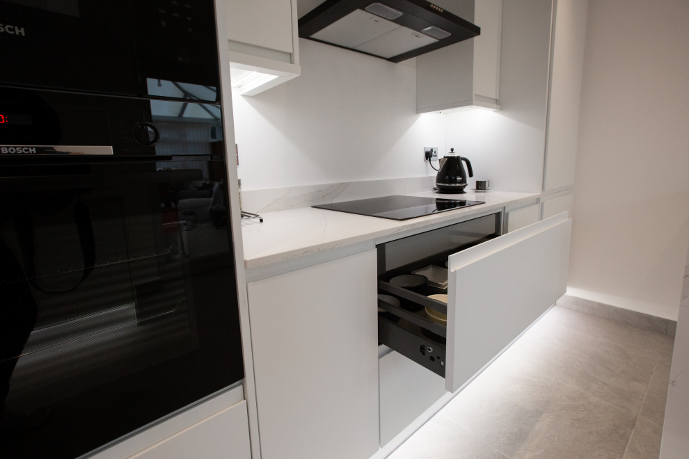 Photo of a medium sized modern galley enclosed kitchen in Manchester with a submerged sink, flat-panel cabinets, white cabinets, quartz worktops, white splashback, engineered quartz splashback, black appliances, cement flooring, an island, grey floors and white worktops.