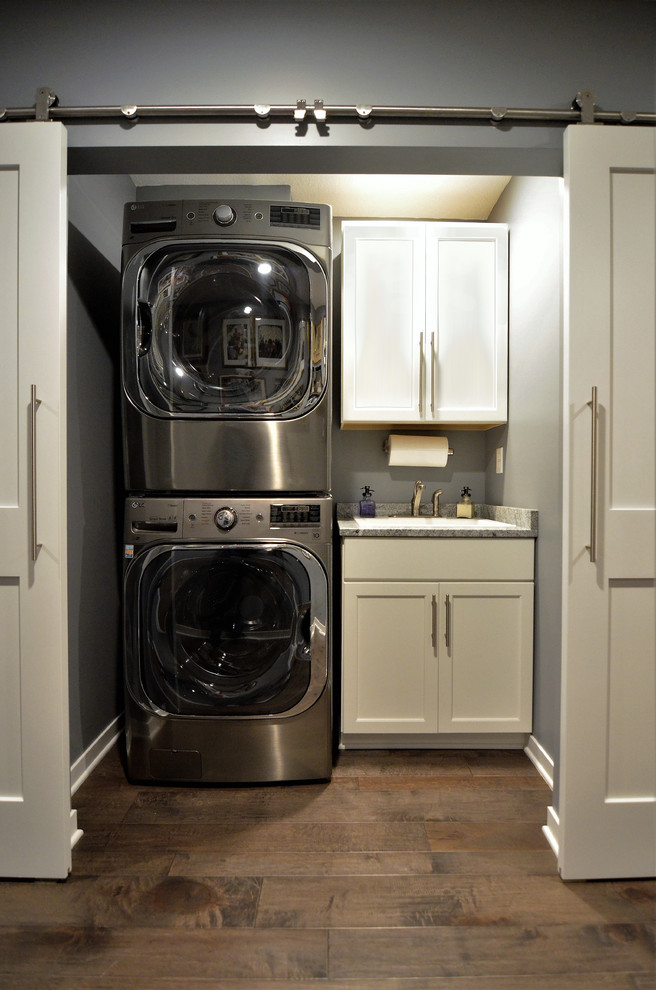 Design ideas for a small transitional single-wall laundry cupboard in Minneapolis with a single-bowl sink, white cabinets, grey walls, medium hardwood floors, a stacked washer and dryer, brown floor and shaker cabinets.