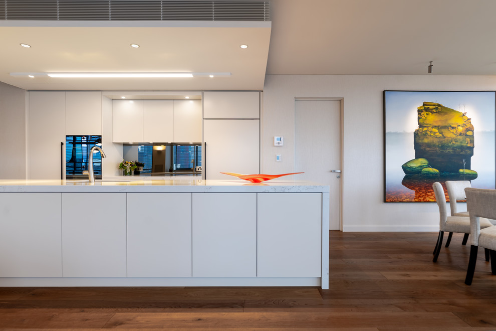 Photo of a contemporary galley eat-in kitchen in Sydney with an undermount sink, flat-panel cabinets, white cabinets, mirror splashback, panelled appliances, medium hardwood floors, with island, brown floor and white benchtop.