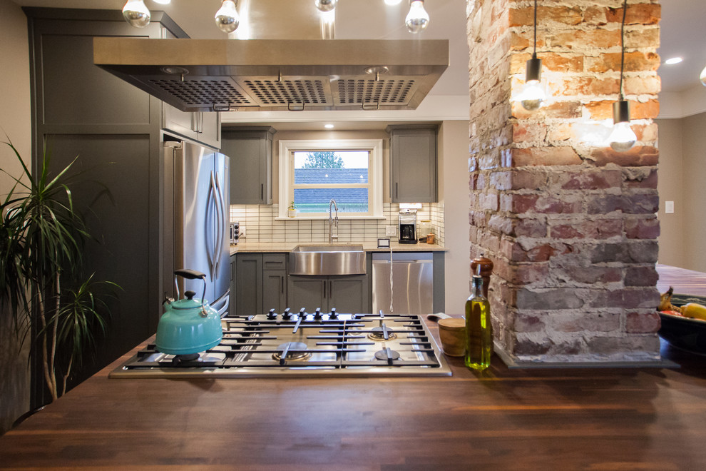 Photo of a small classic l-shaped open plan kitchen in Seattle with a belfast sink, shaker cabinets, grey cabinets, stainless steel appliances, dark hardwood flooring, an island, brown floors, granite worktops, white splashback, porcelain splashback and beige worktops.