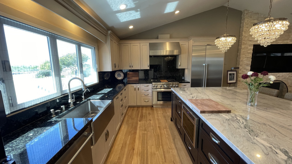 Mid-sized transitional l-shaped kitchen pantry in Orange County with a farmhouse sink, shaker cabinets, white cabinets, granite benchtops, black splashback, granite splashback, stainless steel appliances, light hardwood floors, with island, brown floor, multi-coloured benchtop and coffered.