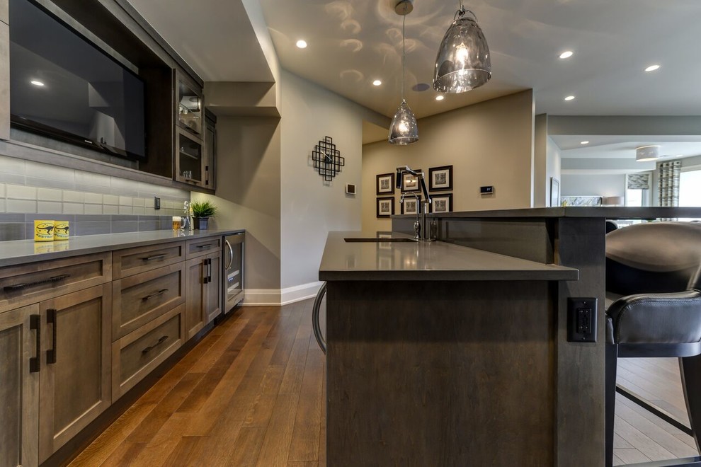 Large transitional l-shaped seated home bar in Edmonton with grey splashback, medium hardwood floors, glass tile splashback, an undermount sink, shaker cabinets, distressed cabinets, solid surface benchtops and brown floor.