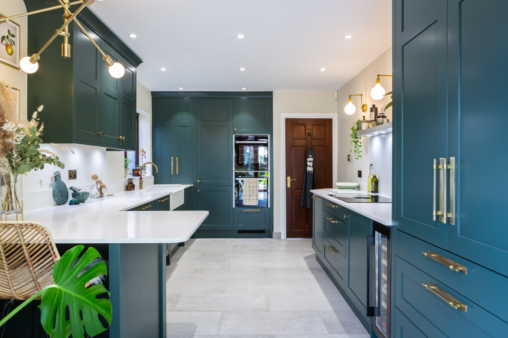 This is an example of a traditional grey and teal u-shaped kitchen in Essex with a belfast sink, shaker cabinets, blue cabinets, a breakfast bar, grey floors and white worktops.