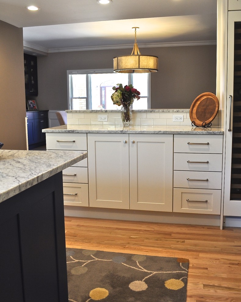Example of a mid-sized transitional l-shaped light wood floor and brown floor enclosed kitchen design in Denver with a farmhouse sink, shaker cabinets, white backsplash, ceramic backsplash, an island, white cabinets, marble countertops and stainless steel appliances