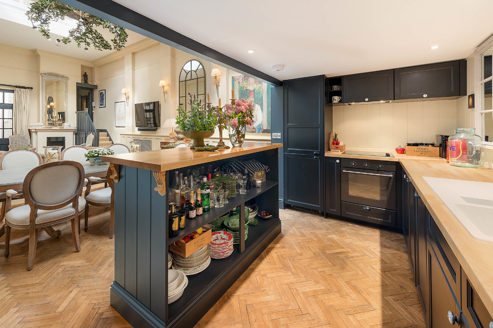 Mid-sized transitional l-shaped kitchen in London with shaker cabinets, blue cabinets, wood benchtops, beige splashback, black appliances, light hardwood floors, with island, brown floor and brown benchtop.
