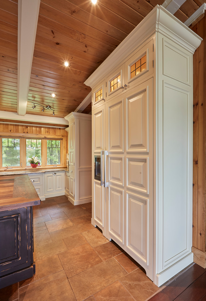 Photo of a large country u-shaped kitchen pantry in Other with a farmhouse sink, raised-panel cabinets, grey cabinets, wood benchtops, beige splashback, travertine splashback, panelled appliances, porcelain floors, with island and brown floor.