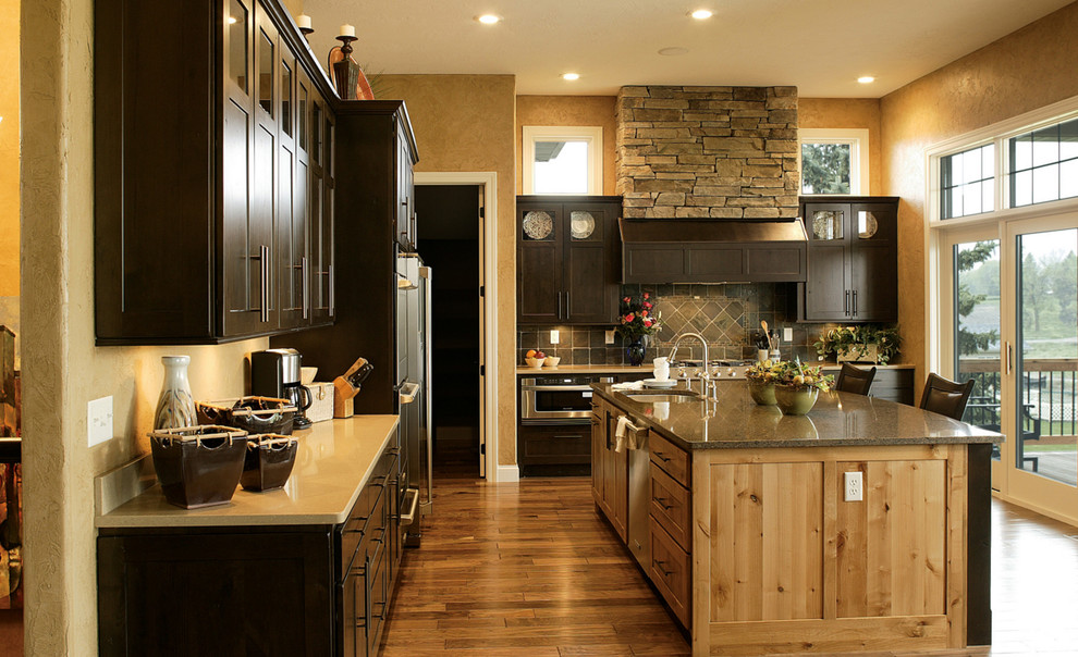Mid-sized mountain style l-shaped medium tone wood floor and brown floor enclosed kitchen photo in Chicago with an undermount sink, recessed-panel cabinets, dark wood cabinets, quartz countertops, gray backsplash, stone tile backsplash, stainless steel appliances and an island