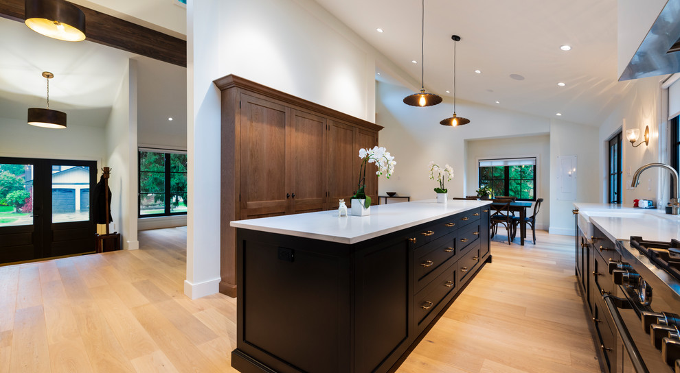 Mid-sized mountain style l-shaped light wood floor and beige floor open concept kitchen photo in Vancouver with a farmhouse sink, shaker cabinets, black cabinets, quartz countertops, paneled appliances, an island and white countertops