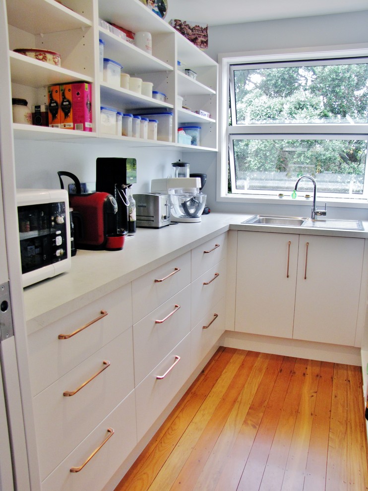 This is an example of a large single-wall eat-in kitchen in Auckland with a triple-bowl sink, raised-panel cabinets, white cabinets, granite benchtops, white splashback, metal splashback, stainless steel appliances, medium hardwood floors, with island, brown floor and white benchtop.