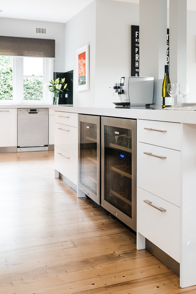 Mid-sized contemporary l-shaped eat-in kitchen in Melbourne with flat-panel cabinets, white cabinets, quartz benchtops, mirror splashback, stainless steel appliances, with island, an undermount sink and light hardwood floors.