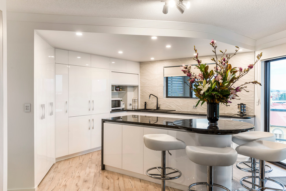Photo of a mid-sized contemporary l-shaped eat-in kitchen in Brisbane with a drop-in sink, flat-panel cabinets, white cabinets, granite benchtops, beige splashback, cement tile splashback, stainless steel appliances, light hardwood floors and with island.