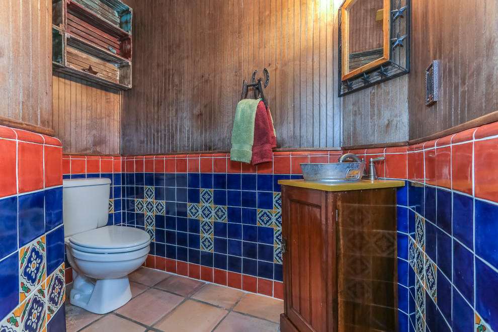 Photo of a large powder room in Houston with a two-piece toilet, brown tile, blue tile, orange tile, multi-coloured tile, porcelain tile, porcelain floors, an undermount sink, granite benchtops, medium wood cabinets, multi-coloured walls, furniture-like cabinets and yellow benchtops.