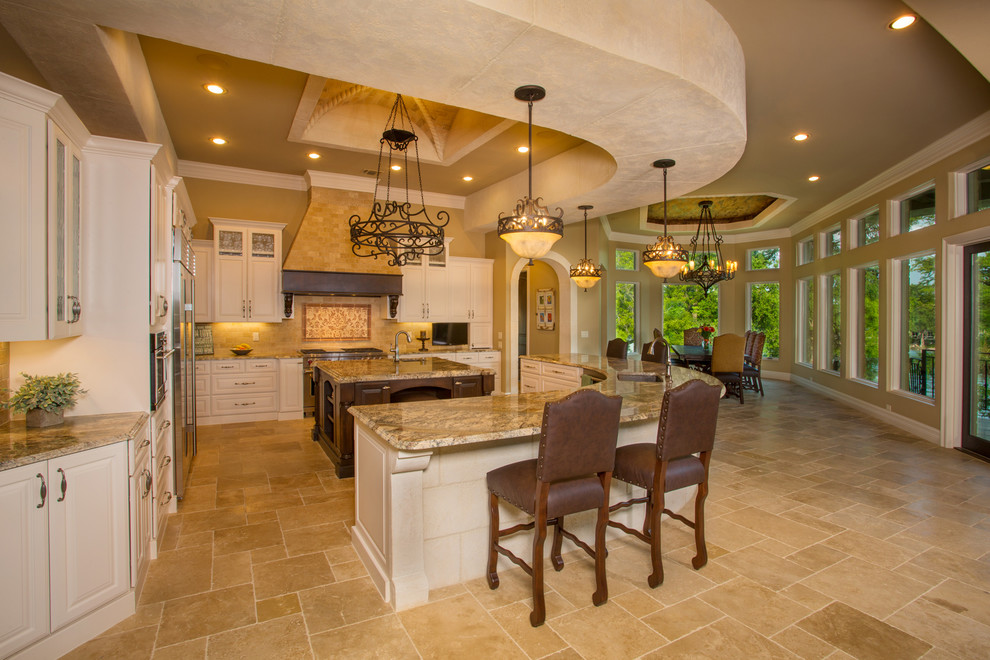 Photo of a large mediterranean u-shaped eat-in kitchen in Austin with an undermount sink, raised-panel cabinets, white cabinets, granite benchtops, beige splashback, stone tile splashback, stainless steel appliances, travertine floors and multiple islands.