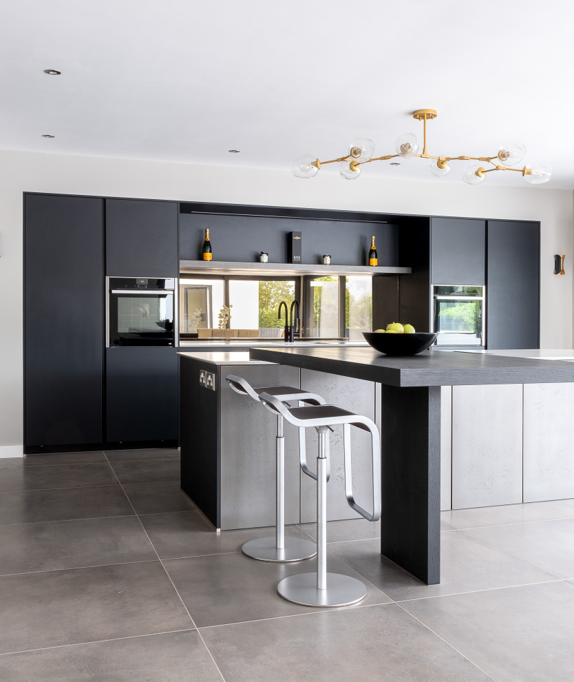 Design ideas for a large contemporary kitchen/diner in Gloucestershire with an integrated sink, flat-panel cabinets, quartz worktops, metallic splashback, mirror splashback, black appliances, an island, grey floors and white worktops.