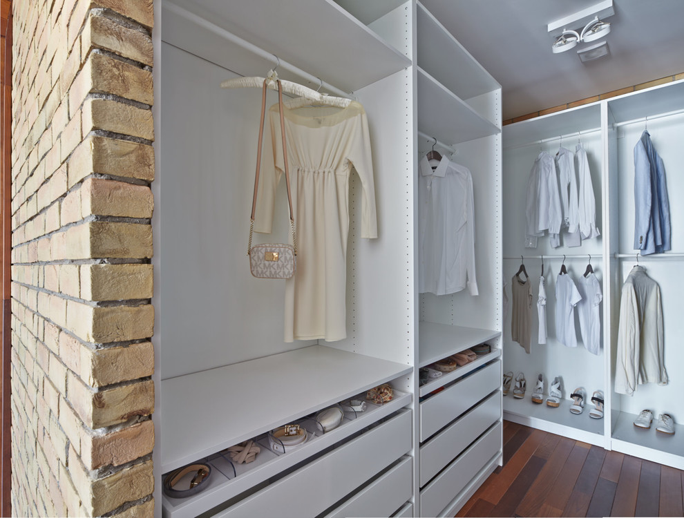 Photo of a contemporary gender-neutral walk-in wardrobe in Moscow with white cabinets, dark hardwood floors, brown floor and open cabinets.
