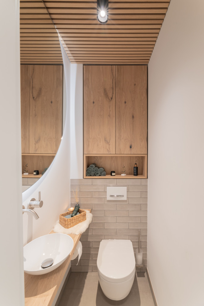Foto på ett litet minimalistiskt beige toalett, med beige skåp, en toalettstol med separat cisternkåpa, grå kakel, glasskiva, vita väggar, klinkergolv i keramik, ett fristående handfat, träbänkskiva och grått golv