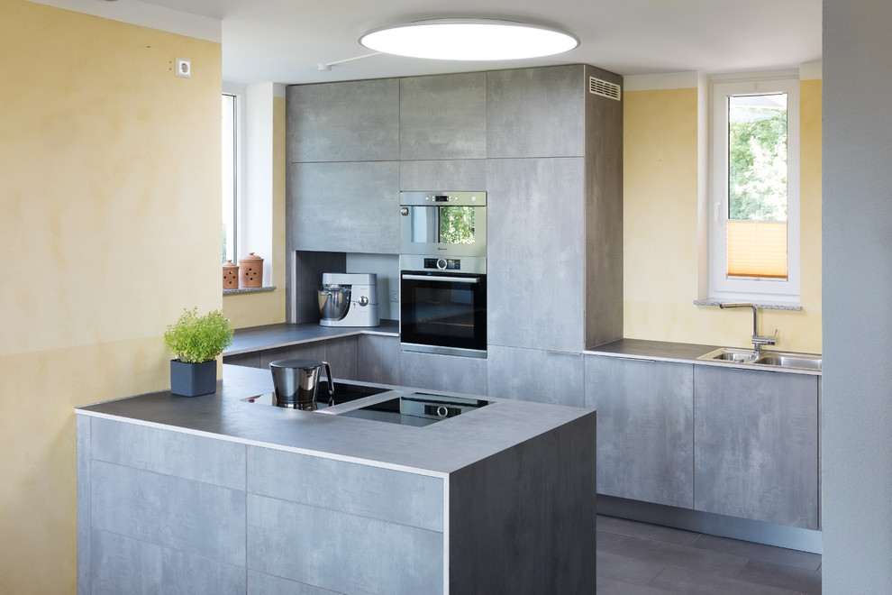This is an example of a mid-sized contemporary u-shaped open plan kitchen in Stuttgart with a double-bowl sink, grey cabinets, laminate benchtops, a peninsula, grey benchtop, flat-panel cabinets, black appliances, grey floor and yellow splashback.