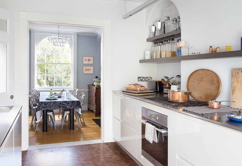 Mid-sized contemporary galley eat-in kitchen in London with stainless steel benchtops, no island, an integrated sink, glass-front cabinets, grey cabinets, white splashback, stainless steel appliances, brown floor and grey benchtop.