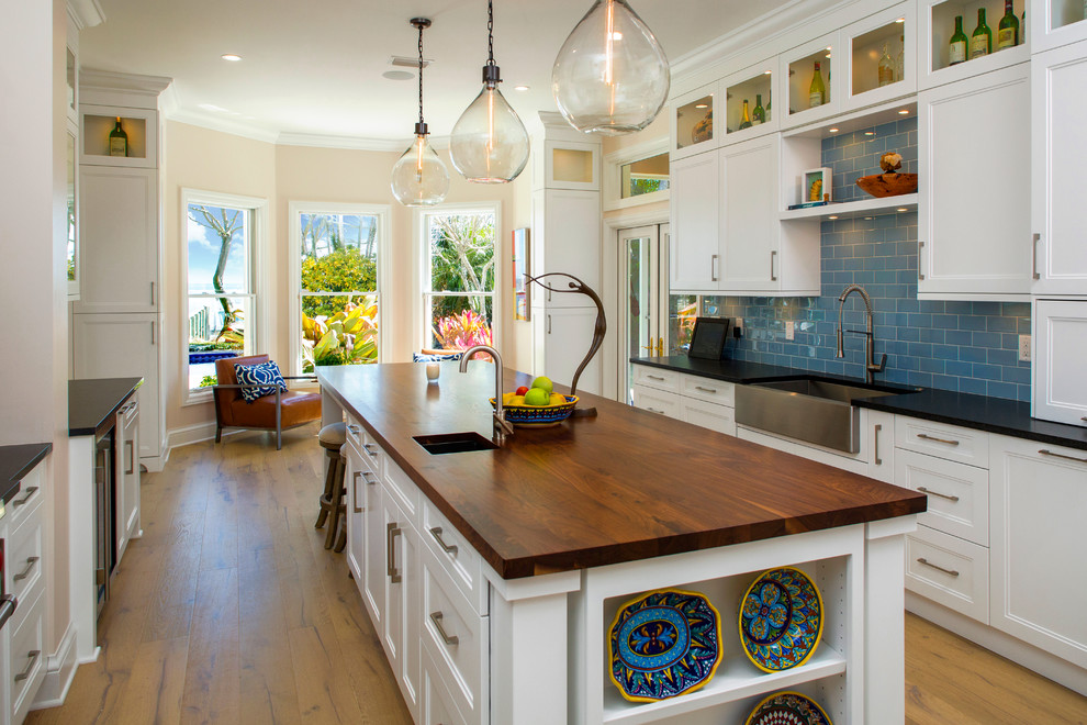 Photo of a large transitional galley eat-in kitchen in Tampa with a farmhouse sink, recessed-panel cabinets, white cabinets, blue splashback, stainless steel appliances, medium hardwood floors, with island, wood benchtops, subway tile splashback, brown floor and black benchtop.