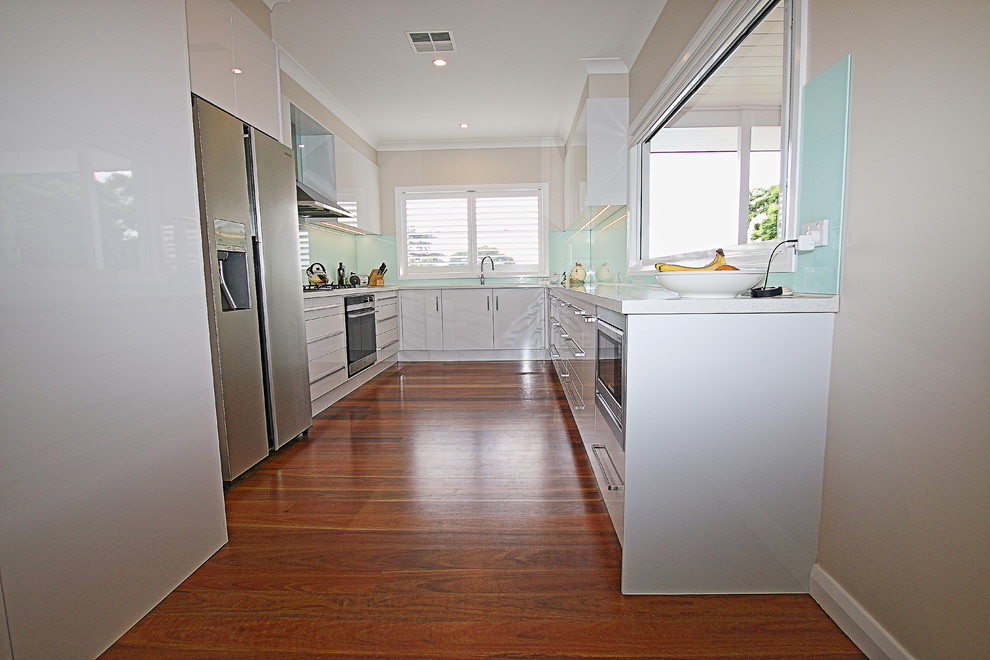 Inspiration for a large contemporary u-shaped kitchen pantry in Sydney with a drop-in sink, flat-panel cabinets, white cabinets, solid surface benchtops, blue splashback, glass sheet splashback, stainless steel appliances, medium hardwood floors and no island.