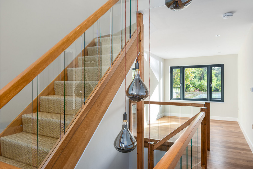 This is an example of a large contemporary carpeted l-shaped staircase in Surrey with carpet risers and glass railing.