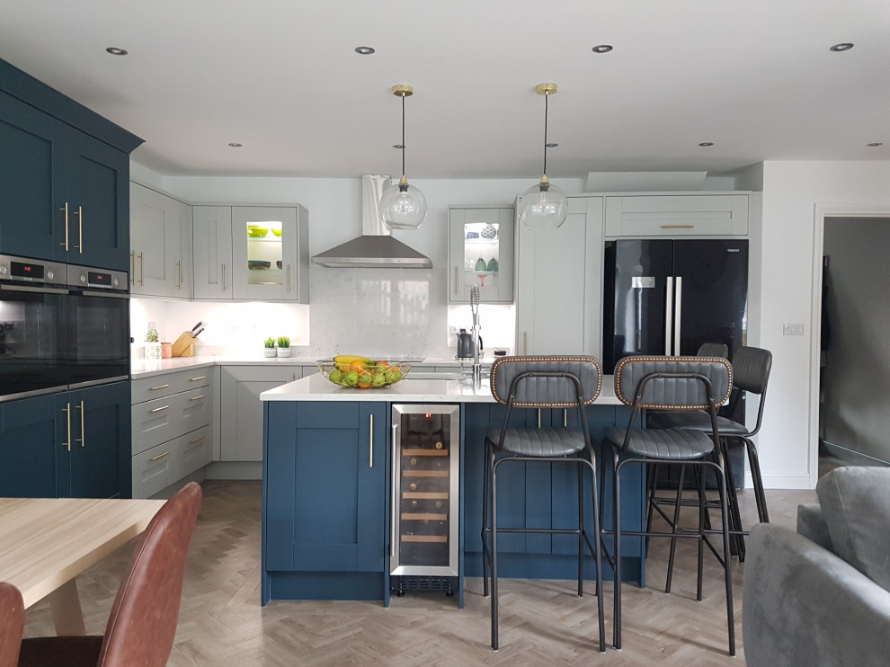 Mid-sized contemporary l-shaped open plan kitchen in West Midlands with an integrated sink, shaker cabinets, blue cabinets, quartzite benchtops, grey splashback, ceramic splashback, black appliances, medium hardwood floors, with island, brown floor and grey benchtop.