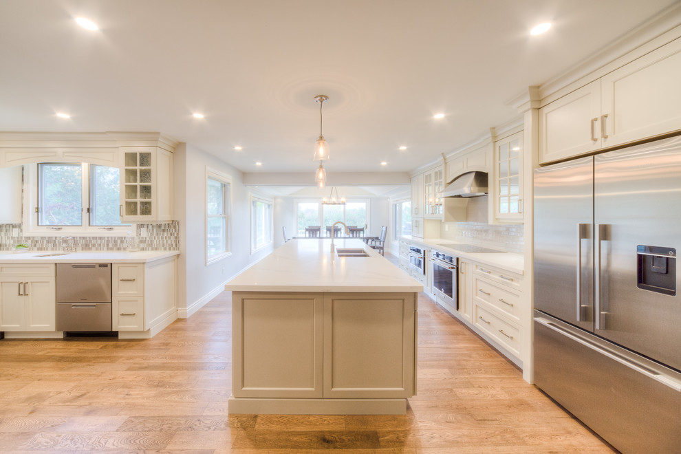 Inspiration for a mid-sized transitional galley light wood floor and beige floor open concept kitchen remodel in Toronto with a double-bowl sink, recessed-panel cabinets, white cabinets, solid surface countertops, white backsplash, porcelain backsplash, stainless steel appliances, an island and white countertops