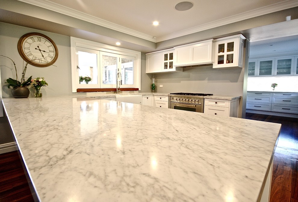 Expansive transitional u-shaped open plan kitchen in Sydney with a farmhouse sink, shaker cabinets, white cabinets, marble benchtops, grey splashback, glass sheet splashback, stainless steel appliances and medium hardwood floors.