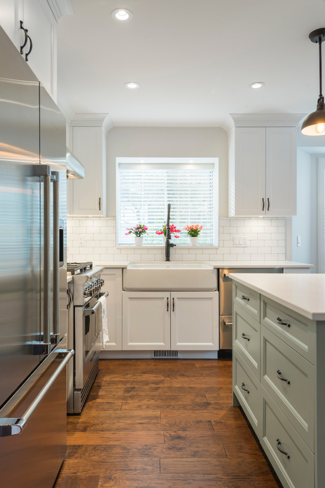 Photo of an expansive traditional l-shaped eat-in kitchen in Vancouver with a farmhouse sink, shaker cabinets, white cabinets, quartz benchtops, white splashback, subway tile splashback, stainless steel appliances, medium hardwood floors, with island, brown floor and white benchtop.