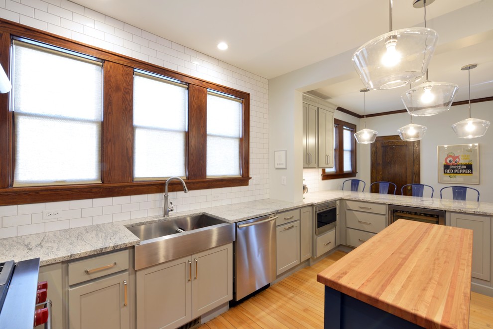 Mid-sized transitional light wood floor and brown floor enclosed kitchen photo with a farmhouse sink, shaker cabinets, gray cabinets, wood countertops, white backsplash, porcelain backsplash, stainless steel appliances, an island and brown countertops