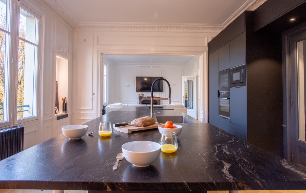 Inspiration for a large contemporary galley open plan kitchen in Rennes with an integrated sink, beaded cabinets, black cabinets, stainless steel worktops, black appliances, light hardwood flooring and an island.