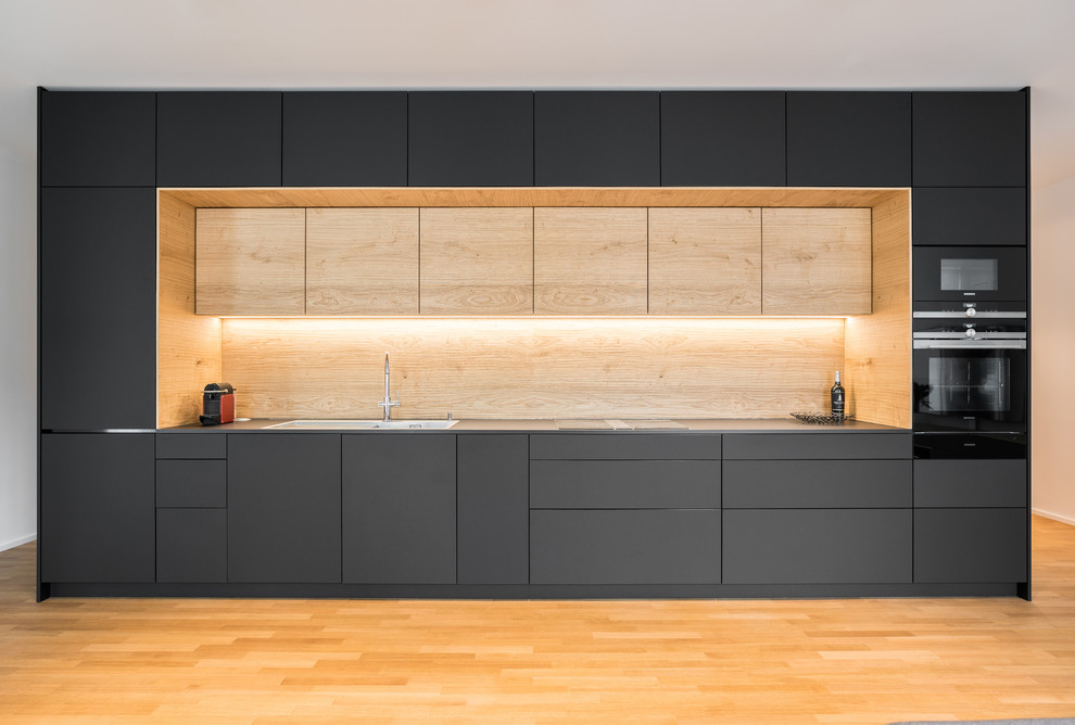 Inspiration for a mid-sized contemporary single-wall open plan kitchen in Stuttgart with a double-bowl sink, flat-panel cabinets, black cabinets, brown splashback, timber splashback, black appliances, medium hardwood floors, no island, brown floor and black benchtop.