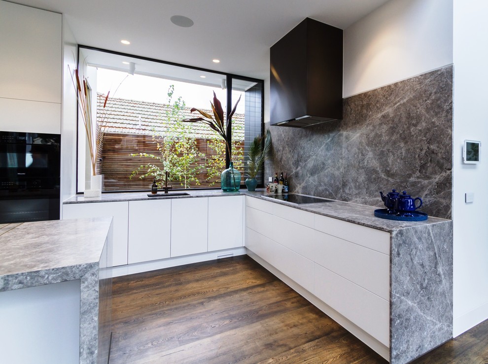 Inspiration for a large modern l-shaped open plan kitchen in Melbourne with an undermount sink, flat-panel cabinets, white cabinets, grey splashback, marble splashback, black appliances, medium hardwood floors, with island, brown floor, grey benchtop and limestone benchtops.