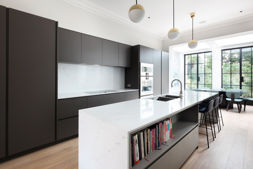 Contemporary single-wall eat-in kitchen in Buckinghamshire with an integrated sink, flat-panel cabinets, black cabinets, quartzite benchtops, white splashback, marble splashback, black appliances, medium hardwood floors, with island, brown floor and white benchtop.